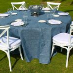 mesa redonda con mantel azul, platos, cubertería y silla crossback en jardín