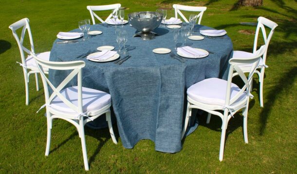 mesa redonda con mantel azul, platos, cubertería y silla crossback en jardín